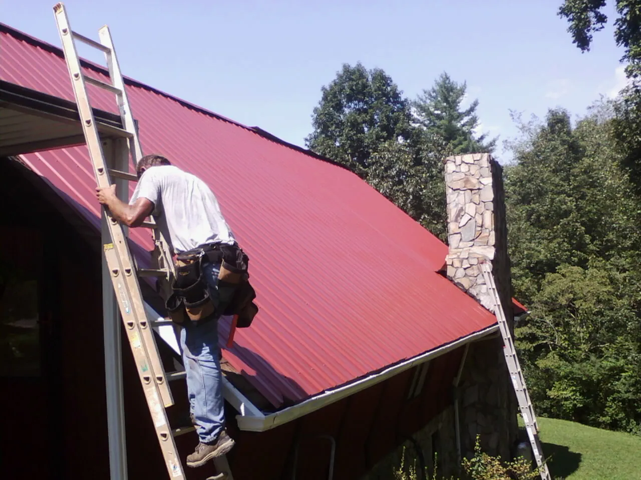 Roofer on a ladder beside a red metal roof and stone chimney — Soffit & Fascia Repair by Roofing Brothers in Summerdale